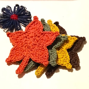 May include: A collection of handmade crochet leaves in autumn colors: orange, green, yellow, and brown. A blue and red decorative flower is also present. These items are likely for fall decor or craft projects.