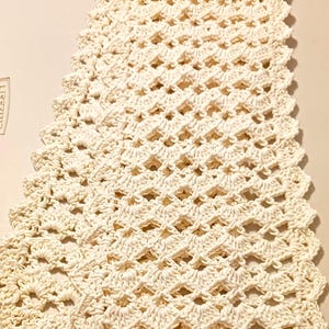 May include: A rectangular off-white crochet table runner featuring a repeating shell stitch pattern. The textile has an openwork design with scalloped edges along the sides and ends, creating a textured, lacy appearance throughout the entire piece.