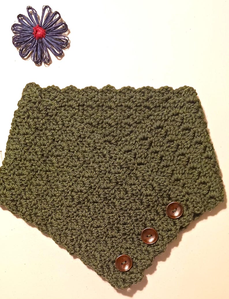 May include: An olive green crocheted cowl with three brown buttons. The cowl features a textured stitch pattern and a scalloped edge. A blue and red decorative flower is in the upper left corner. The item is likely handmade.