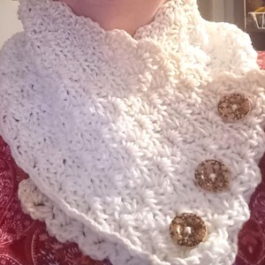 May include: A cream-coloured, crocheted cowl with a scalloped edge and three decorative, round buttons. The handmade neck warmer is a cosy accessory. The buttons are a light brown colour.