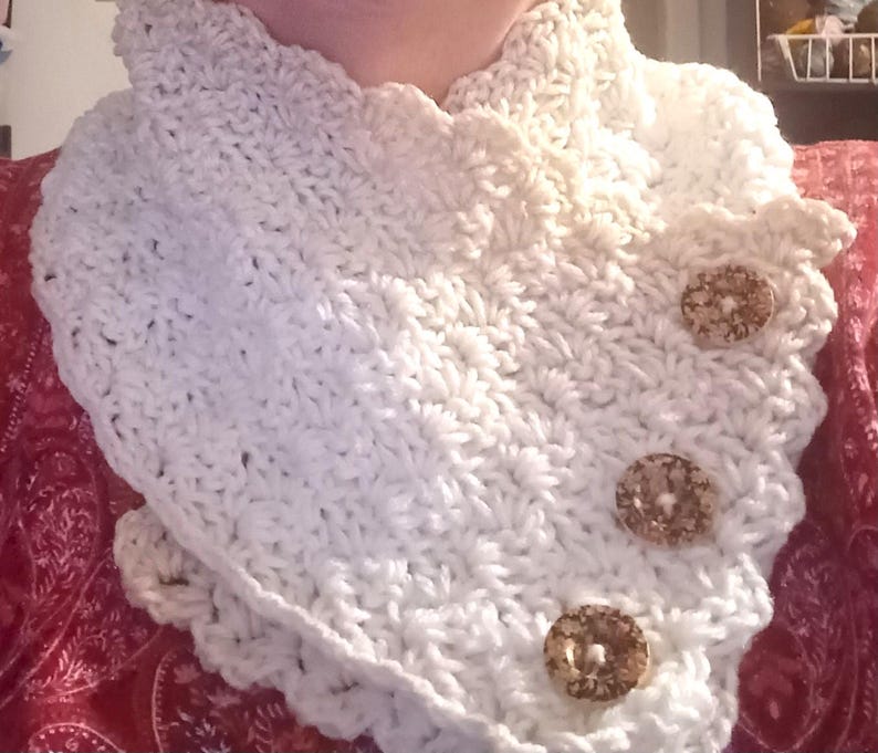 May include: A cream-coloured, crocheted cowl with a scalloped edge and three decorative, round buttons. The handmade neck warmer is a cosy accessory. The buttons are a light brown colour.