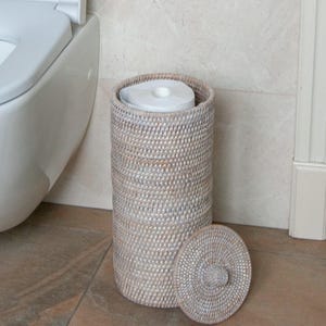 May include: A cylindrical toilet paper holder crafted from woven light brown and white rattan. The tall, open-top container features a matching circular lid with a small central knob, designed to store rolls of toilet paper in a bathroom setting.