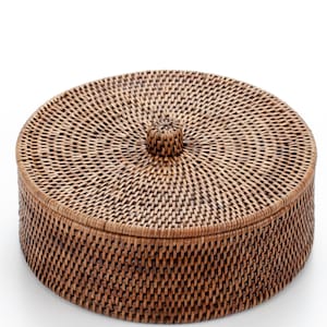 以下が含まれることがあります： 丸い編み込みラタン製の収納ボックス。蓋付き。ボックスは温かみのある茶色で、蓋の上部にノブが付いています。きめ細かい編み込み模様が、テクスチャのある表面を作り出しています。小物の収納に適しています。直径約20cm。