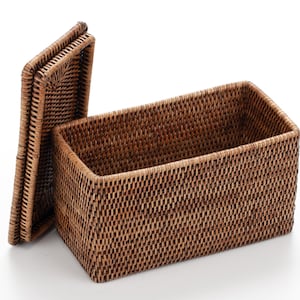 May include: A rectangular woven rattan storage basket with a matching lid. The basket is a warm brown color and has a textured surface. The lid is slightly ajar, revealing the interior of the basket. This decorative basket is ideal for home organization.