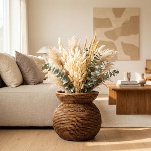 Op de afbeelding: Een bruine, geweven rieten vaas gevuld met gedroogd pampasgras, tarwe en eucalyptusbladeren. De vaas heeft een bolle vorm met een uitlopende bovenkant. De opstelling staat op een houten vloer.