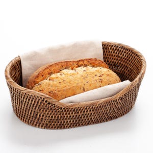 May include: An oval-shaped, woven brown bread basket with two loaves of bread. One loaf is light brown with seeds, and the other is darker. A beige linen cloth lines the basket.