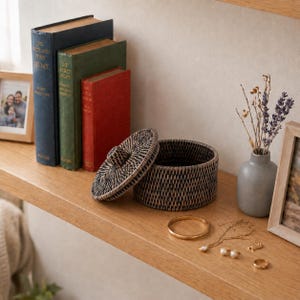Puede incluir: Una caja de almacenamiento redonda de ratán trenzado marrón oscuro con tapa a juego, junto a tres libros de tapa dura en azul, verde y rojo. Un pequeño jarrón gris contiene tallos de lavanda seca y joyas doradas, incluyendo un brazalete.