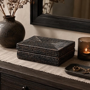May include: A rectangular storage box crafted from dark brown woven rattan material. The box features a hinged lid with a tight, uniform weave pattern and a flat top surface. The structure is compact and low-profile, showcasing a natural, textured finish throughout the exterior of the container.