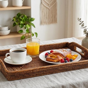 Op de afbeelding: Een rechthoekig dienblad van geweven rotan met handvatten bevat een witte koffiekop en schotel, een glas sinaasappelsap en een bord met gebak en fruit. De dienblad staat op een lichtgrijze tafellaken.