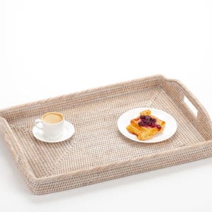 May include: A rectangular, cream-coloured woven tray with handles, holding a white cup of coffee on a saucer and a plate with a pastry. The tray has a textured surface and a light border.