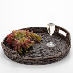 Op de afbeelding: Een donkerbruin geweven dienblad met handvatten, met een tros druiven en een champagnefluit. De druiven zijn een mix van rood, groen en geel. De fluit is gevuld met een lichtgekleurde vloeistof.