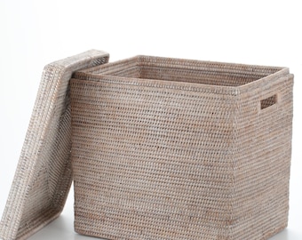 Puede incluir: Una cesta de almacenamiento rectangular tejida en color beige natural. La tapa está ligeramente abierta, revelando el interior. La cesta tiene asas laterales. La cesta está hecha de un material tejido apretado.