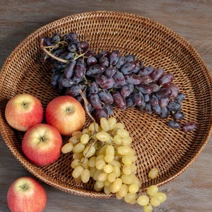 Puede incluir: Una bandeja redonda de mimbre tejido llena de fruta fresca. La bandeja contiene un racimo de uvas moradas oscuras, un racimo de uvas verdes y tres manzanas rojas. La bandeja está sobre una superficie de madera.
