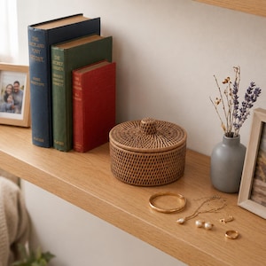 Puede incluir: Una caja de almacenamiento redonda de ratán trenzado con tapa se encuentra sobre un estante de madera junto a tres libros de tapa dura en azul, verde y rojo. Cerca, un pequeño jarrón gris contiene lavanda seca, un brazalete, un collar y anillos.