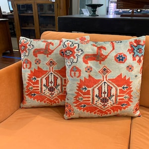 May include: Two square throw pillows with a light blue background and a repeating pattern in red, orange, and blue. The pattern includes stylized deer and geometric shapes. The pillows are on an orange sofa.