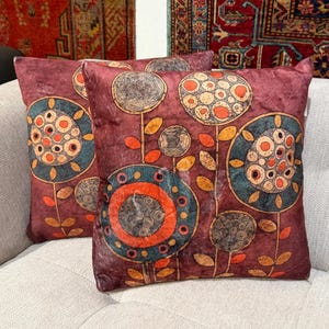 May include: Two decorative throw pillows with a floral design. The pillows are a deep burgundy color with a pattern of stylized flowers in teal, orange, and gold. The pillows are placed on a light-colored sofa.