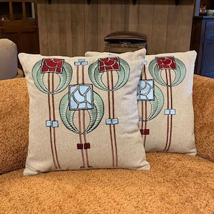 May include: Two square throw pillows with a beige background and an embroidered floral design. The design features red, green, and blue geometric shapes and lines. The pillows are placed on an orange sofa.