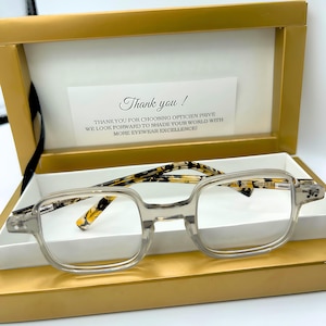 Peut inclure: Lunettes à monture transparente de forme carrée, avec des branches à motifs, présentées dans un coffret doré. Le coffret a un intérieur blanc et une note de remerciement.