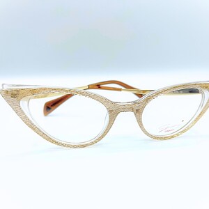 Peut inclure: Lunettes œil de chat avec une monture dorée texturée et des verres transparents. Les lunettes ont un fin pont et des branches dorées, et les verres sont de forme ovale. Le nom de la marque est imprimé sur le verre droit.