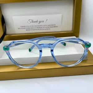 Peut inclure: Lunettes rondes bleu clair avec des accents verts sur les branches. Les lunettes sont dans une boîte avec un mot de remerciement.