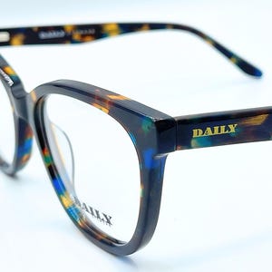 May include: Eyeglasses with a tortoiseshell pattern in blue, yellow, and brown. The frames have a cat-eye shape and the word "DAILY" is printed on the temple and lens. The glasses are set against a white background.