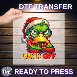 May include: DTF transfer with a cartoon duck wearing a Santa hat, glasses, and a red jacket. The duck has an angry expression and the words "DUCK OFF" are below it. The text "DTF TRANSFER" and "READY TO PRESS" are also present.