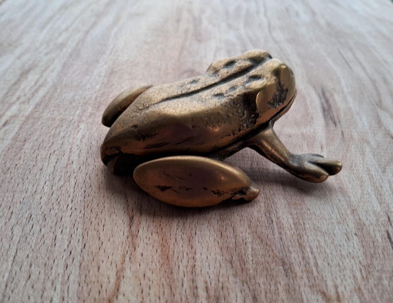 May include: A small, gold-colored frog figurine made of metal. The frog is sitting on a wooden surface.