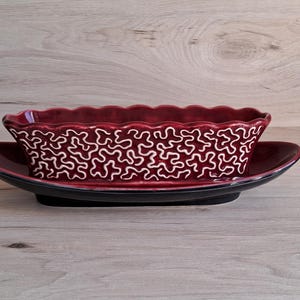 May include: A red ceramic planter with a white, abstract pattern. The planter has a scalloped edge and sits on a black saucer.