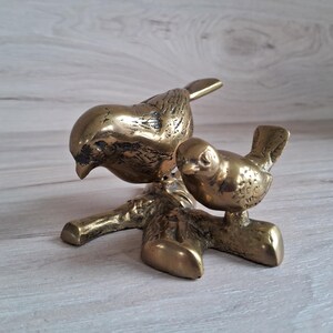 May include: A gold-toned metal sculpture of two birds perched on a branch. The larger bird is facing down, while the smaller bird is facing forward. The sculpture is detailed with textured feathers and a realistic branch.