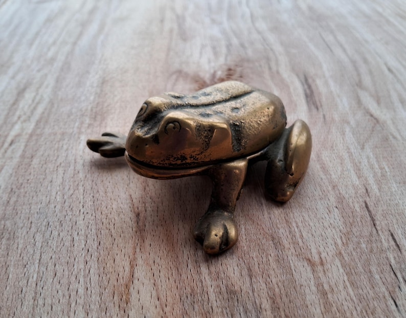 May include: A brass frog figurine with a detailed design. The frog is sitting on a wooden surface.