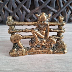 May include: A brass figurine depicting a seated fairy figure on a mushroom, with a small fence behind it. The fairy has pointed ears and is seated with its arms raised. The figurine is detailed and has a vintage appearance.
