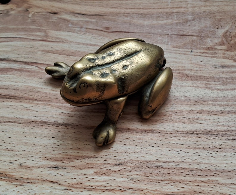 May include: A brass frog figurine with a detailed textured surface. The frog is sitting on a wooden surface.