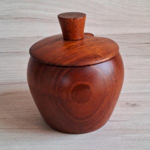 May include: A wooden jar with a lid, made of dark brown wood with a smooth, polished finish. The jar has a rounded shape and a slightly flared top.