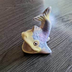 Może przedstawiać: Ceramiczna figurka ryby w odcieniach żółci, błękitu i fioletu. Ryba ma otwartą paszczę i uniesiony ogon. Figurka spoczywa na drewnianej powierzchni.