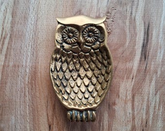 May include: A brass owl figurine with detailed feather and eye patterns. The owl is a golden color and is set against a light brown wooden surface. The owl has a small beak and feet.
