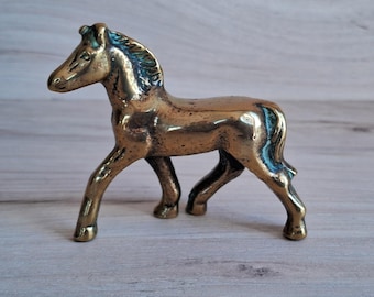 May include: A small, gold-colored brass horse figurine. The horse is standing with its head turned to the left and its legs slightly bent. The figurine is detailed with a mane and tail.