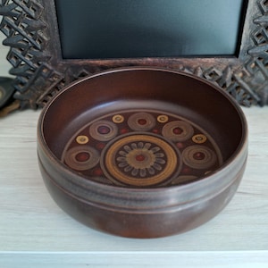May include: A brown ceramic bowl with a decorative pattern in the center. The pattern features concentric circles in shades of brown, tan, and red. The bowl has a smooth, rounded shape and a dark brown exterior.