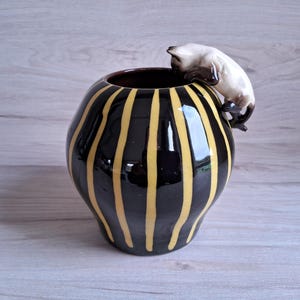 Könnte beinhalten: Eine schwarze und gelbe gestreifte Keramikvase mit einer weißen Keramikkatzenfigur auf dem Rand.