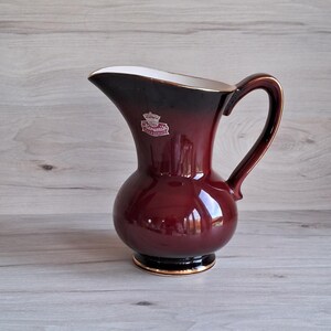 May include: A dark red ceramic pitcher with a gold rim and a white interior. The pitcher has a rounded body and a curved handle. The logo "Keramik" with a crown is visible on the front.