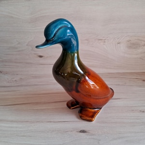 Puede incluir: Figura de pato de cerámica con acabado brillante. El pato tiene cabeza y cuello de color turquesa, que transicionan a un cuerpo marrón y naranja. La figura está en posición de pie, mostrando sus detalles.