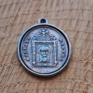 Op de afbeelding: Zilverkleurige religieuze hanger met de woorden "SAGRADO ROSTRO" rondom de rand. De hanger heeft een centraal beeld van een bebaard gezicht in een rechthoekig kader. De hanger heeft een lus aan de bovenkant om op te hangen.