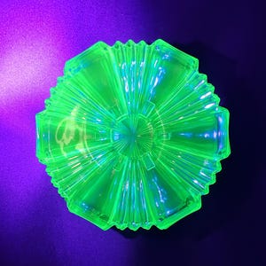 May include: A bright green, hexagonal glass bowl with a fluted design. The bowl features a starburst pattern in the centre and a scalloped edge. The glass glows against a purple and black background, possibly uranium glass.