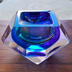 May include: A faceted glass bowl featuring a deep blue and purple gradient with a clear, angular exterior. The interior displays a smooth, rounded basin that transitions into sharp, geometric edges, creating a reflective and multi-dimensional appearance.