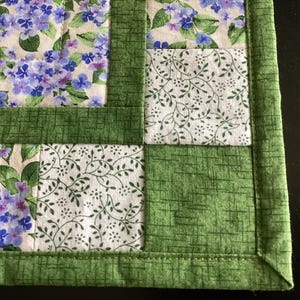 Puede incluir: Un mantel individual acolchado con un dise&ntilde;o floral. El mantel individual presenta cuadrados de tela con flores de hortensia azules y moradas, tela blanca con hojas verdes y bordes verdes. Los bordes est&aacute;n cuidadosamente cosidos.