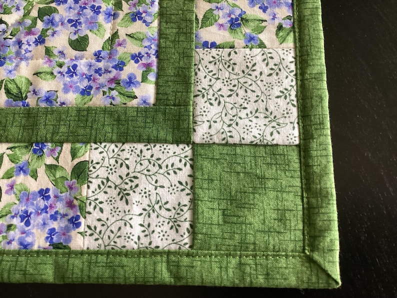Puede incluir: Un mantel individual acolchado con un dise&ntilde;o floral. El mantel individual presenta cuadrados de tela con flores de hortensia azules y moradas, tela blanca con hojas verdes y bordes verdes. Los bordes est&aacute;n cuidadosamente cosidos.