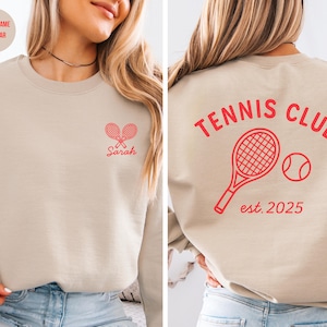 May include: Beige sweatshirt with red tennis-themed graphics. The front features crossed rackets and the name "Sarah." The back displays "TENNIS CLUB" above a racket and ball, with "est. 2025" below. A casual, sporty design.