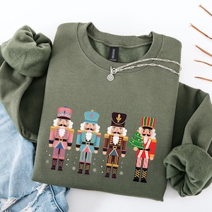 Könnte beinhalten: Ein olivgrünes Sweatshirt mit einem bunten Aufdruck von vier Nussknackern. Die Nussknacker tragen rosa, blaue, braune und rote Uniformen mit Schneeflocken-Akzenten. Eine silberne Halskette liegt obenauf.