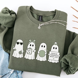 May include: A military green crewneck sweatshirt featuring a graphic of four white ghosts. The ghosts are depicted with physical therapy items including a dumbbell, a clipboard labeled PT, a green exercise ball, and a sign reading PHYSICAL THERAPISTS MOVE LIVES.