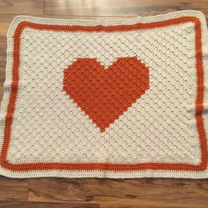 May include: A crocheted blanket with a large orange heart in the center. The blanket has a white background and an orange border.