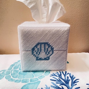 May include: A white tissue box with a blue seashell design. The box is made of a textured material and has a diagonal pattern. White tissues are pulled from the top. The box sits on a white towel with a blue sea turtle and coral design.